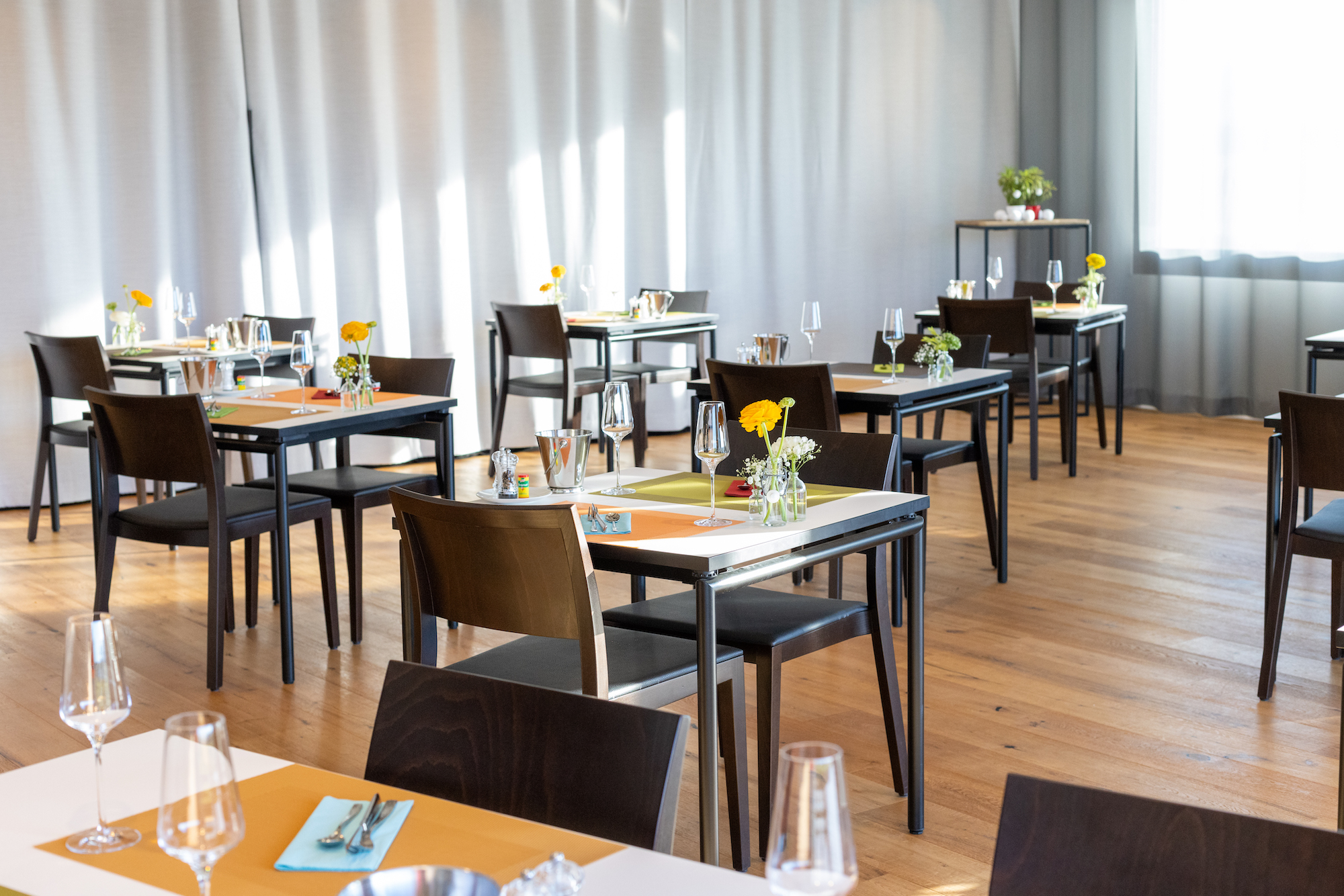 Ein heller Raum mit mehreren gedeckten Tischen für einen Brunch. Jeder Tisch hat Gläser, Blumen und farbige Tischsets. Dunkle Stühle stehen um die Tische. Im Hintergrund helle Vorhänge und ein Holzboden.